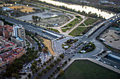 Cars navigate the busy roundabout in Seville while the sun sets, casting a warm glow over the city landscape and river.