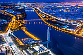 Glowing lights illuminate the Guadalquivir River in Seville, showcasing the Schildler Tower and several notable bridges at night.