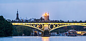 Bridge illuminated at twilight with Torre del Oro visible against the Seville skyline.