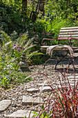 Sitzplatz mit Steinmännchen vor Beet mit Schnittlauch, Chinaschilf, Lampenputzergras und Farnblätter im sommerlichen Garten 
