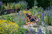 Frau im Liegestuhl zwischen Rittersporn (Delphinium) und Färberkamille (Anthemis tinctoria) im sommerlichen Garten