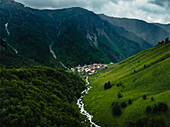 Luftaufnahme eines malerischen Dorfes in einem üppig grünen Tal mit einem gewundenen Fluss und majestätischen Bergen, Adishi, Georgien