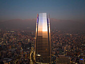 Luftaufnahme des Sonnenuntergangs, der sich in einem hohen Wolkenkratzer spiegelt, mit Stadtbild und Andengebirge in der Ferne, Santiago, Chile
