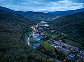 Luftaufnahme einer malerischen Stadt in einem ruhigen Tal, umgeben von üppigen Wäldern und kurvenreichen Straßen bei Sonnenuntergang, Thann, Haut-Rhin, Frankreich
