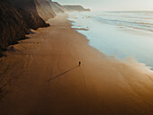 Luftaufnahme des ruhigen Strandes Praia do Cordoama mit einer Person, die bei Sonnenuntergang am Sandstrand spazieren geht, Vila do Bispo, Portugal