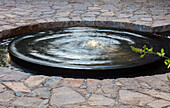 Runder, schwarzer Springbrunnen im Terrassenboden aus Steinplatten
