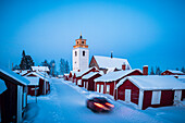 Auto fährt auf verschneiter Straße zwischen traditionellen roten Häusern in der Altstadt von Gammelstad in der Winterdämmerung, Lulea, Schweden