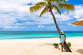 Woman in turquoise dress leaning on palm tree on idyllic tropical beach, Barbuda, Antigua and Barbuda, Caribbean, West Indies
