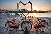 Toscolano maderno gulf, with position for touristic photos, red bike with heart, church of St. Andrea, boats, with sun at sunrise, Lake of Garda, Toscolano Maderno, Brescia province, Lombardy, Italy, north Italy, Europe, south Europe