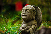 A joyful Warabe-Jizo statue cradling a child, decorated with a red maple leaf, amidst vibrant autumn grass and leaves, Kyoto, Honshu, Japan