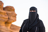 Woman in a niqab at Face Rock, a sandstone rock formation in shape of human head profile, Hegra, UNESCO, AlUla, Medina Province, Saudi Arabia
