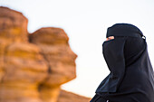 Woman in a niqab at Face Rock, a sandstone rock formation in shape of human head profile, Hegra, UNESCO, AlUla, Medina Province, Saudi Arabia