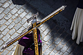 Hooded penitent holding golden adorned cross during Holy Week procession in Seville, Spain.