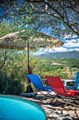 Relaxing poolside view featuring colorful lounge chairs and a straw umbrella amidst nature