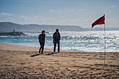 Couple walking on Nazare beach with red flag, Portugal