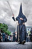 In Seville during Holy Week, a Nazareno in traditional robes holds a long wax candle while walking solemnly among the crowd.