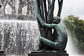 A bronze sculpture depicts a naked figure joyfully hugging a tree in Vigeland Park, surrounded by a backdrop of a large fountain in Oslo.