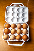 A white egg container holding twelve brown eggs placed atop a wooden table surface.