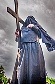 A hooded penitent solemnly carries a wooden cross in Seville, celebrating the deeply rooted traditions of Holy Week in Andalusia.