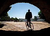 A cyclist's silhouette beneath a bridge arch in Seville with sunny scenery.