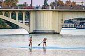 Sevilla, Spanien, 8. Oktober 2019, Zwei Mädchen paddeln beim Surfen auf dem Fluss Guadalquivir in Sevilla, mit der San-Telmo-Brücke als atemberaubende Kulisse für den Sonnenuntergang.