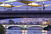 Kanufahrer befahren den Fluss Guadalquivir, während die Brücke Cristo de la Expiración und die Triana-Brücke den Abendhimmel in Sevilla erhellen.