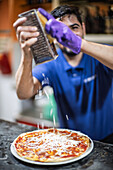 A skilled pizza chef in a bustling kitchen freshly grates Parmesan cheese over a delicious pizza topped with vibrant tomato sauce.