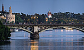 Twilight casts a beautiful glow over Triana Bridge in Seville, highlighting the Torre del Oro and lively atmosphere along the riverbank.