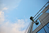 Niedriger Blickwinkel auf einen männlichen Höhenarbeiter, der an einem Glasgebäude unter blauem Himmel hängt