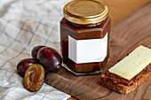 A hexagonal jar of plum compote with an empty label stands on a wooden surface. Fresh plums and a slice of bread with compote and cheese add a rustic touch to the scene.