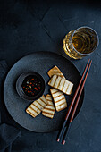 Top view of grilled tofu slices on a black plate with spicy soya sauce and a glass of whiskey. The dark, textured background emphasises the aesthetic, served with wooden chopsticks.
