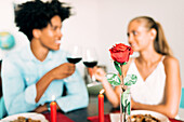 A romantic table setting with a couple toasting with glasses of red wine. A bright red rose in a vase and candles emphasise the intimate atmosphere. Perfect for celebrating a relationship.