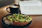 A delicious slice of avocado toast on a black plate, garnished with seeds and greens, next to a steaming cup of coffee on a cosy wooden table that creates a warm morning atmosphere.