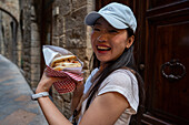 An Asian woman savours traditional Italian street food as she explores the charming stone-paved streets of Volterra in Tuscany. Her indulgence reflects the cultural richness and culinary heritage of Italy