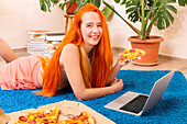 A person with bright red hair relaxes on a blue carpet and enjoys a pizza while using a laptop, surrounded by potted plants and piles of books.