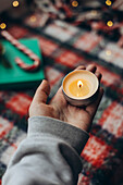 Cropped unrecognisable person holding a lit candle against a festive background with blurred Christmas elements.