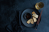 Grilled slices of tofu and soya cheese, served on a black plate with rice and spicy pepper sauce, accompanied by chopsticks and a glass of drink, against a dark slate background.