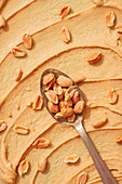 Top view of a soft peanut butter spread with a spoonful of raw, shelled peanuts deeply embedded, providing a rich textural contrast in natural orange tones.