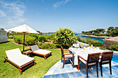 A sun-drenched hotel terrace on the coast with a lush garden, elegant dining area and inviting sun loungers that invite you to relax, overlooking turquoise waters and a peaceful horizon