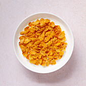 Top view of a white bowl full of crunchy golden cornflakes, isolated on a light-coloured, textured background. Perfect for a healthy and quick breakfast.