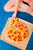 A person sits comfortably on a bright blue carpet and reaches for a slice of pepperoni pizza from an open box.
