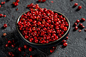 Top view of a dark bowl of bright red peppercorns scattered on a textured dark background, bringing out the vibrant colour and glossy texture of the spice.