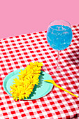 A lively summer picnic scene with a glass of iced blue drink and a plate of yellow flowers on a red chequered tablecloth against a pink background.