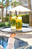 Two colourful cocktails with mint and lime on a tray held by a poolside waiter. The sunny setting with green plants and parasol is the epitome of relaxation.