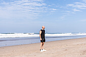 A bald man in black sportswear and white trainers stands pensively on a sandy beach in Bali, gazing at the ocean waves under a clear blue sky that symbolises a cheerful holiday mood.