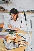 A young blind woman slices cucumbers in a bright, modern kitchen. A loaf of bread lies in a basket next to her. A cane indicates a visual impairment. Inclusive culinary activity.