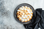A lemon cake with toasted meringue lies on a dark-coloured plate, complemented by a cloth. The cake with its golden tips offers a tantalising sight against a textured background.