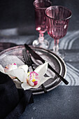 A sophisticated table setting with textured plates, dark-coloured cutlery and delicate white orchids. Two pink glasses emphasise the romantic atmosphere against a soft background.