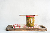 A jar of preserved homemade organic peas is placed on a wooden board and sealed with a red dotted lid. On top is a spoon that evokes a rustic kitchen atmosphere.