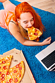 A person with bright red hair enjoys a slice of pepperoni pizza while lounging on a soft blue carpet next to a laptop, capturing a cosy day at home